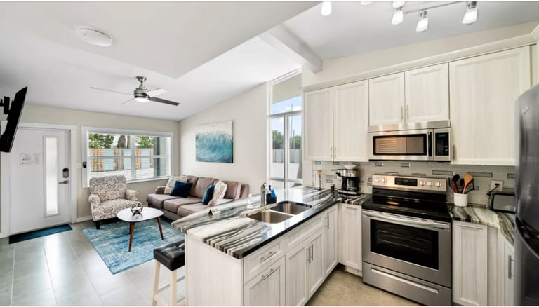 Modern open-concept kitchen and living room with cream cabinets, stainless steel appliances, and beige sofa with windows