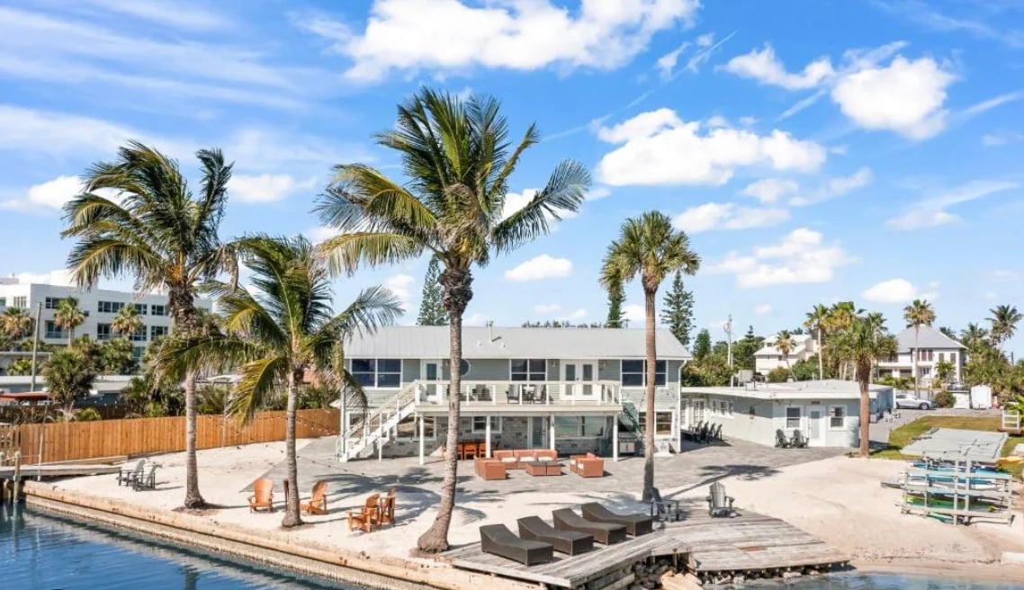 Waterfront beach house with elevated deck, palm trees, and blue sky with white clouds