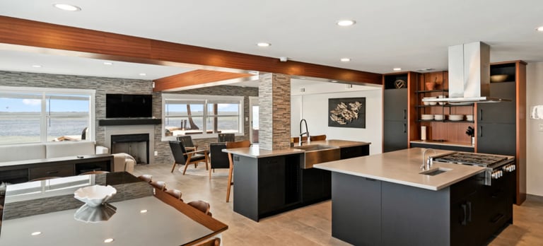 Modern open-concept kitchen and living room with black island, wood beams, fireplace, and ocean view
