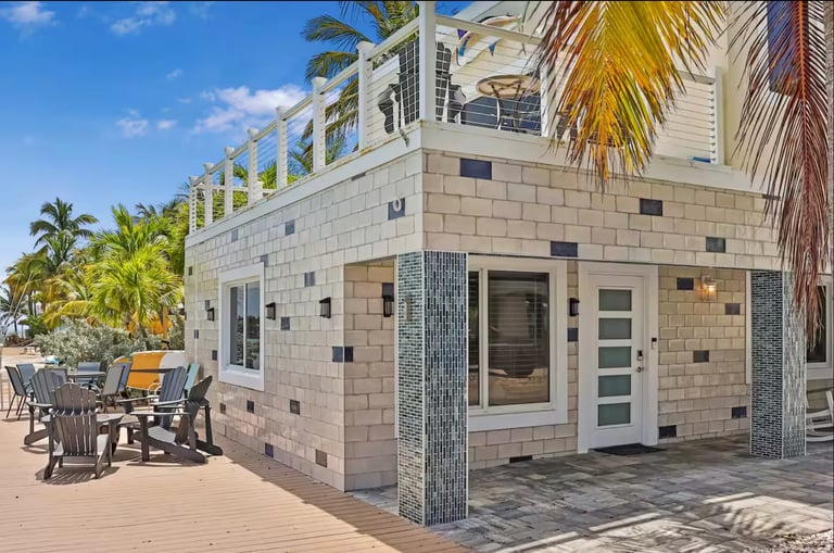 Modern beachfront house with stone exterior, rooftop deck, palm trees, and outdoor seating area on wooden patio